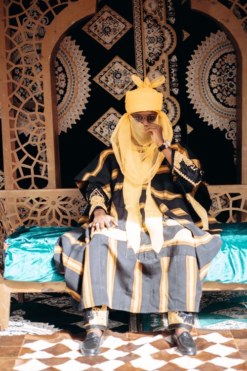 Free Man wearing elaborate Nigerian traditional attire, seated on a decorated chair indoors. Stock Photo
