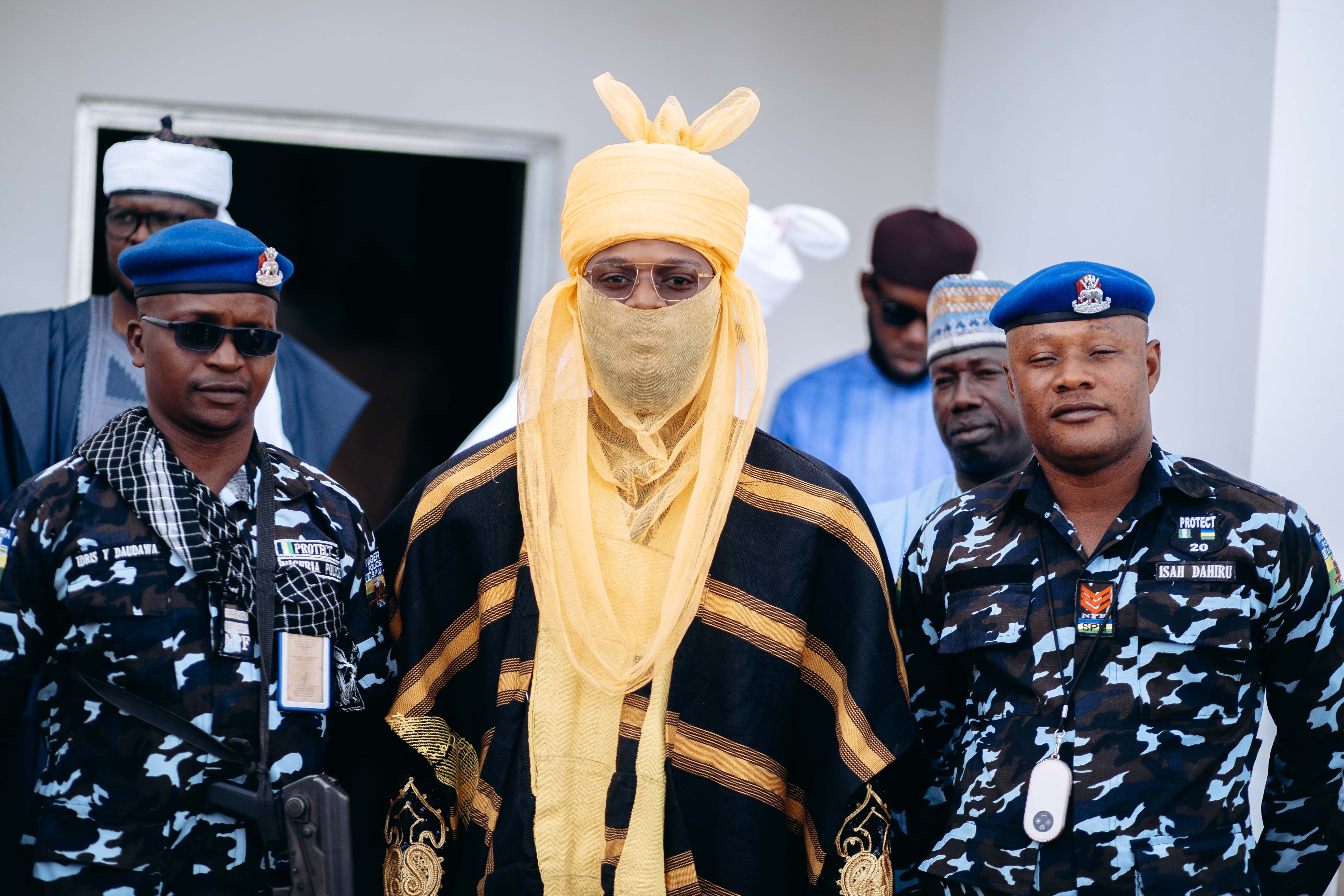 A traditional leader in ceremonial attire stands with a group of security personnel outdoors.