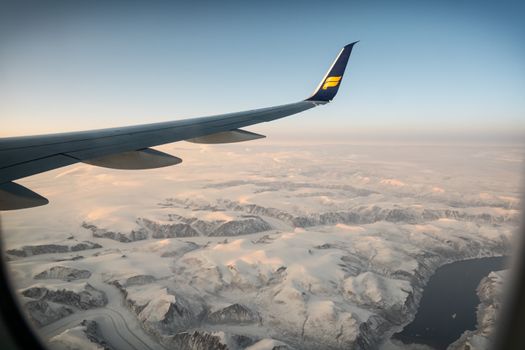 Aerial view from airplane window overlooking icy mountains at sunset, capturing serene winter landscape.