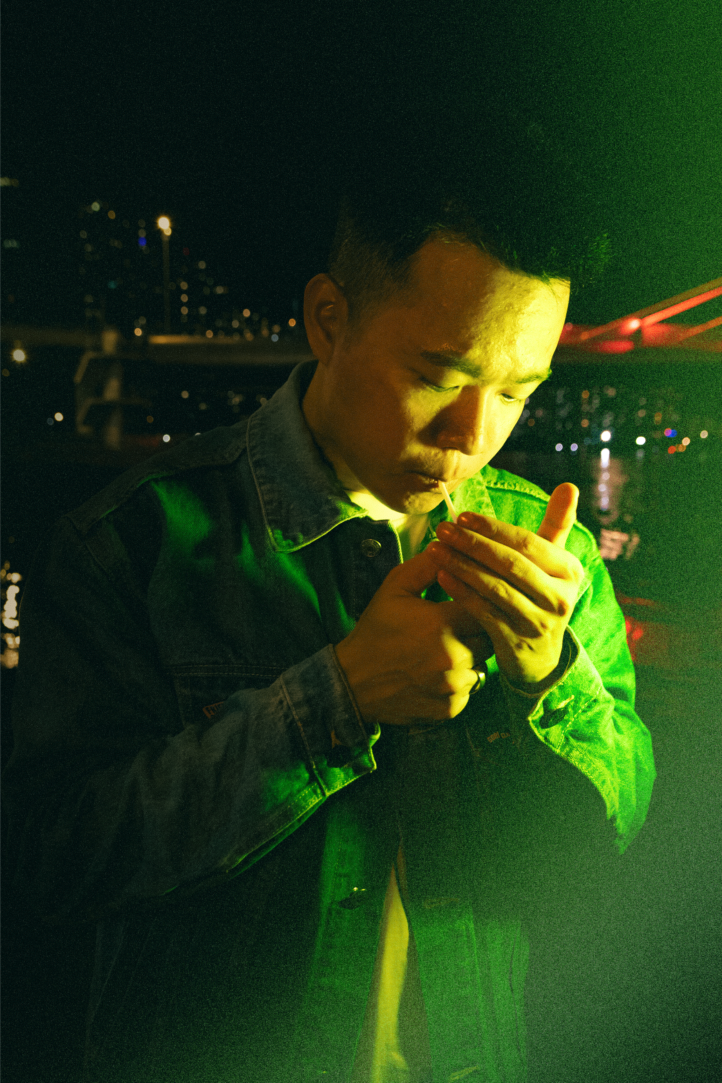 A man in a denim jacket lights a cigarette amidst the neon cityscape of Ho Chi Minh City at night.