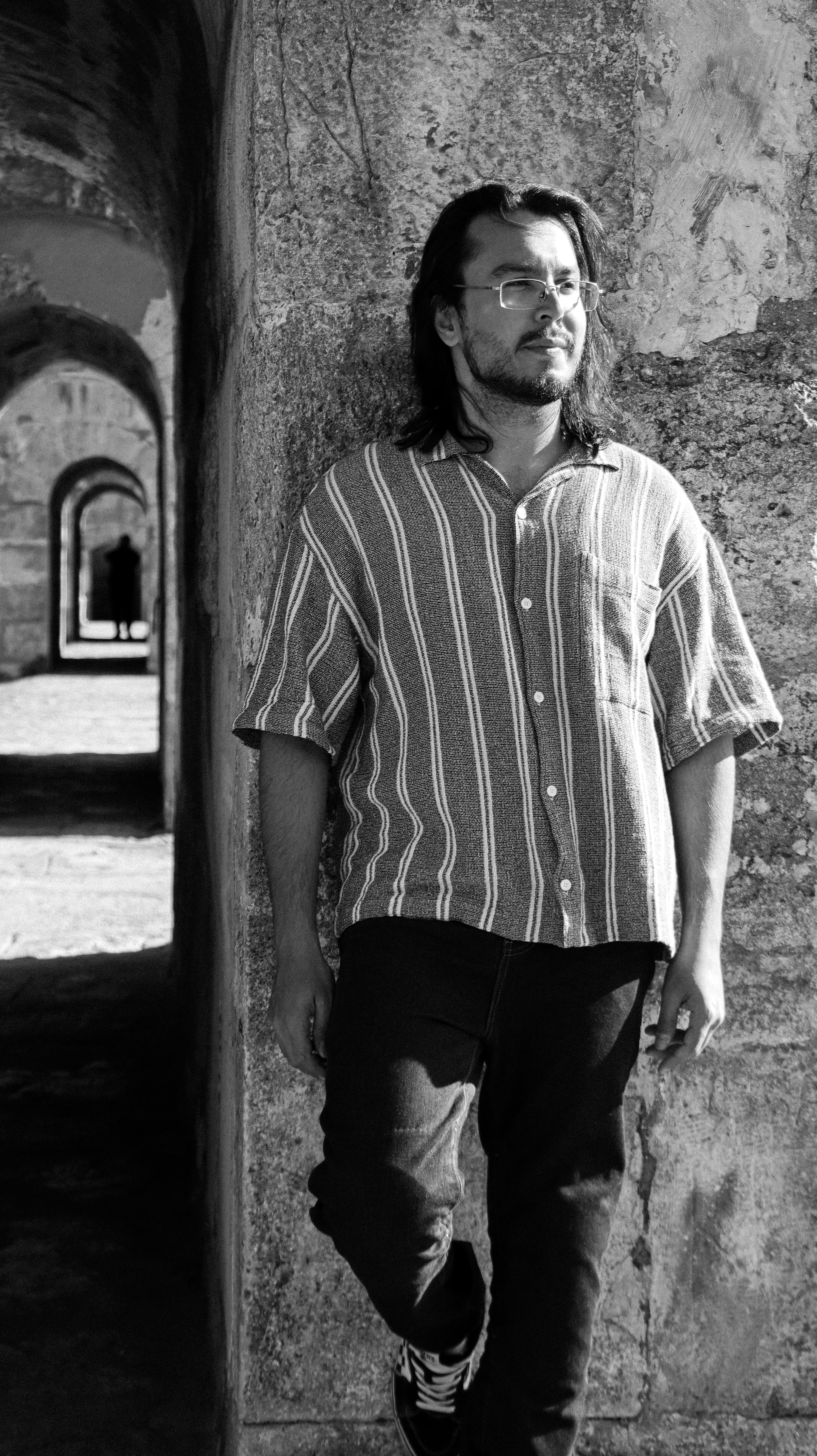 Portrait of a man leaning casually against a historical arched stone wall, evoking a sense of vintage charm.