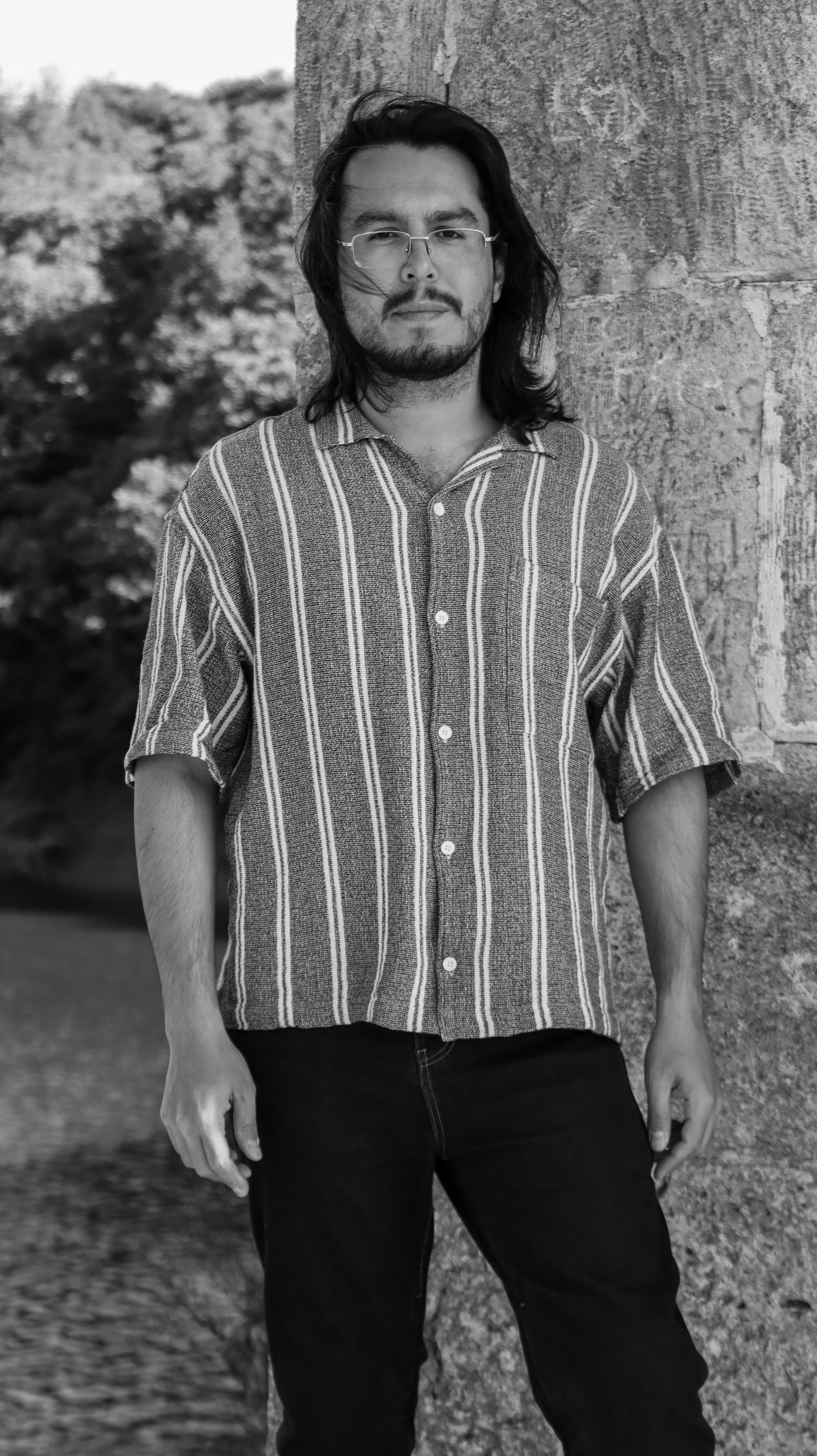Black and white photo of a man in glasses, standing by a tree trunk in casual attire.