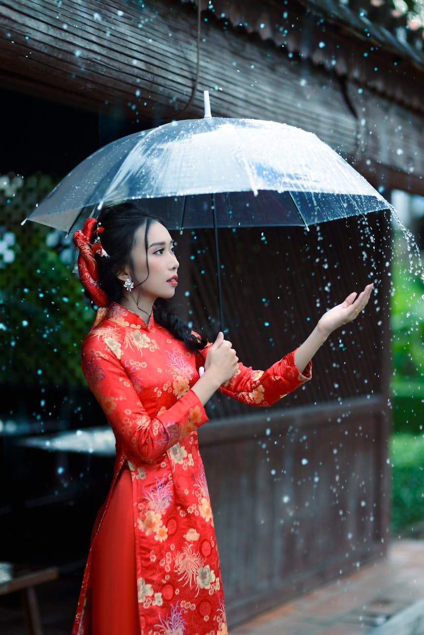 A young woman in red ao dai holds an umbrella during a rain shower.