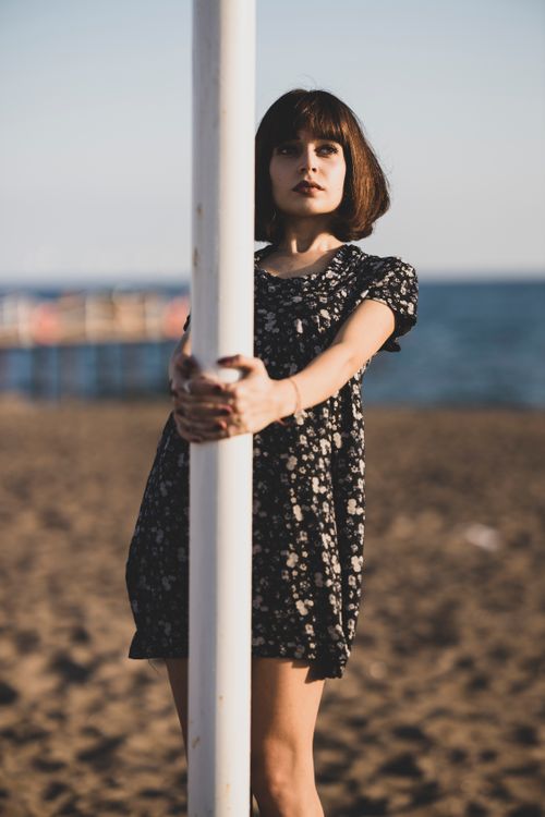 Free A young woman in a floral dress poses by a pole on a sandy beach at sunset. Stock Photo