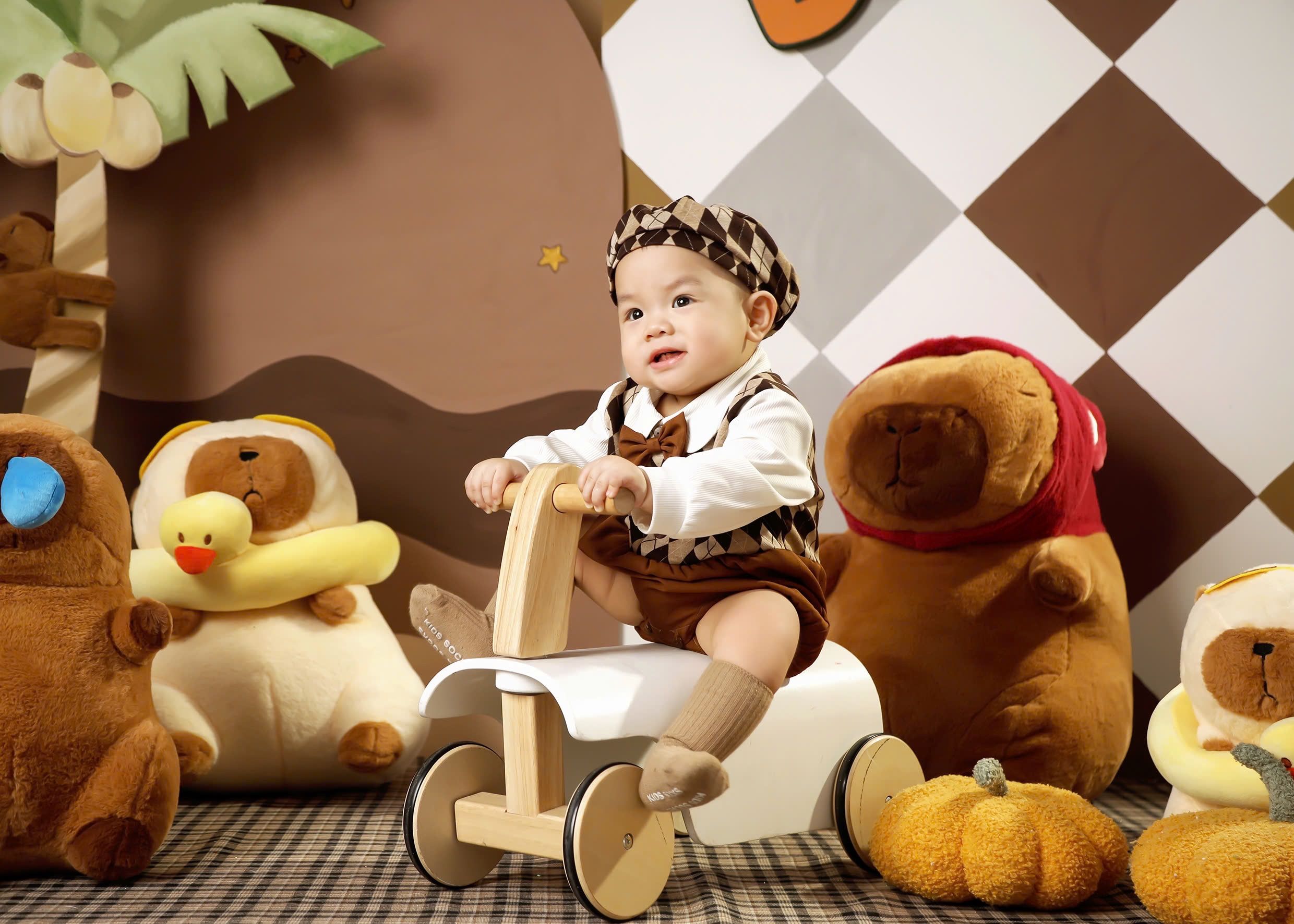 Cute baby sitting on wooden toy surrounded by plush animals and pumpkins in a cozy playroom setting.