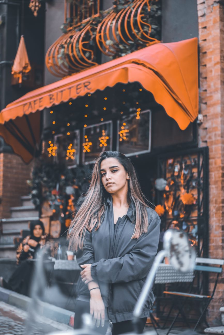 Stylish woman posing outside a chic cafe with a vibrant orange awning, exuding modern fashion vibes.