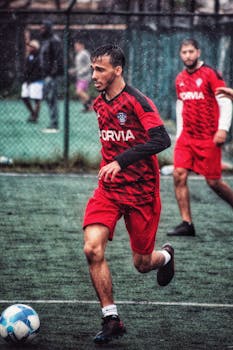 Two men playing soccer in the rain on a wet field, wearing red uniforms.