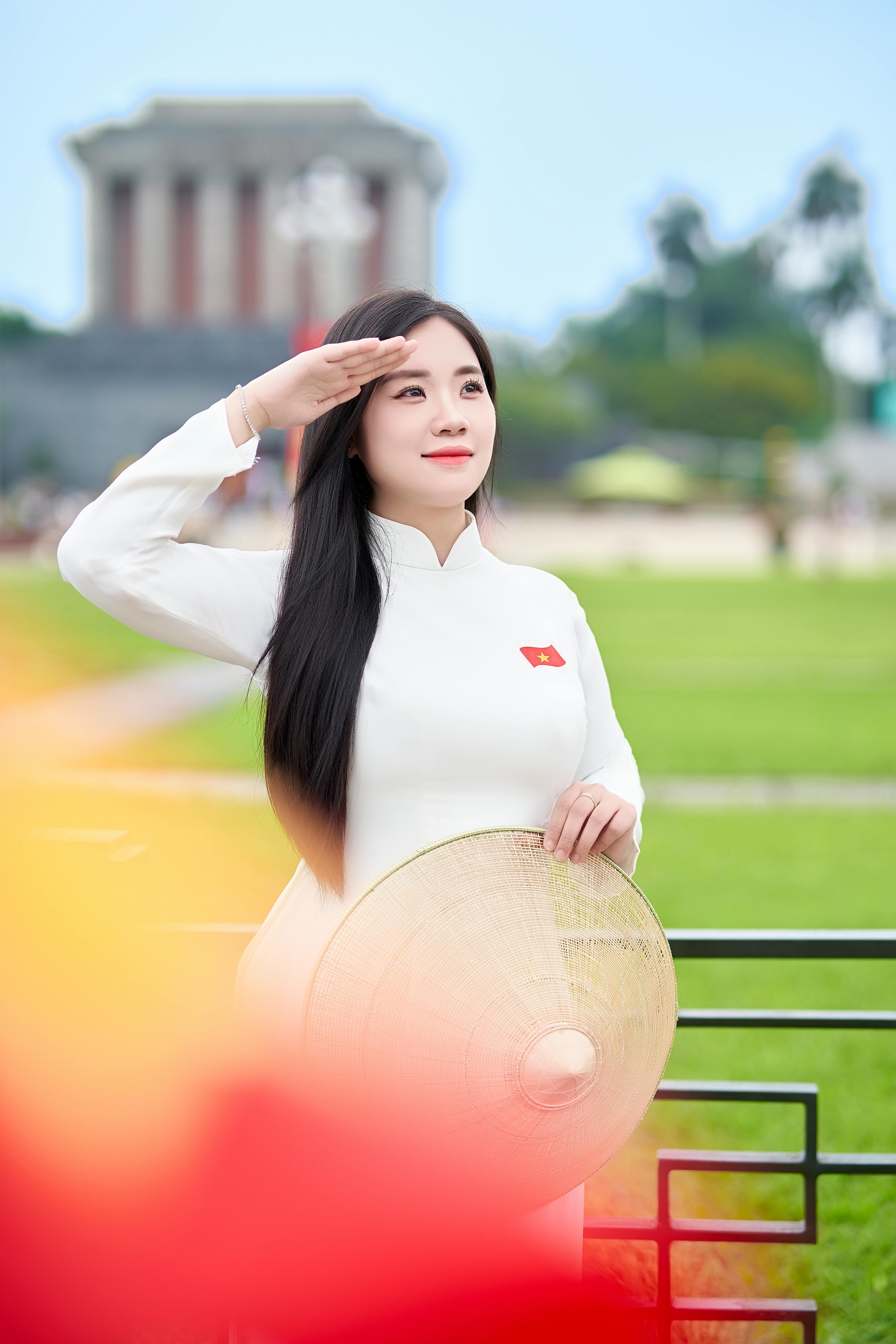 Woman in white áo dài holding conical hat and saluting outside a recognizable Vietnamese landmark.