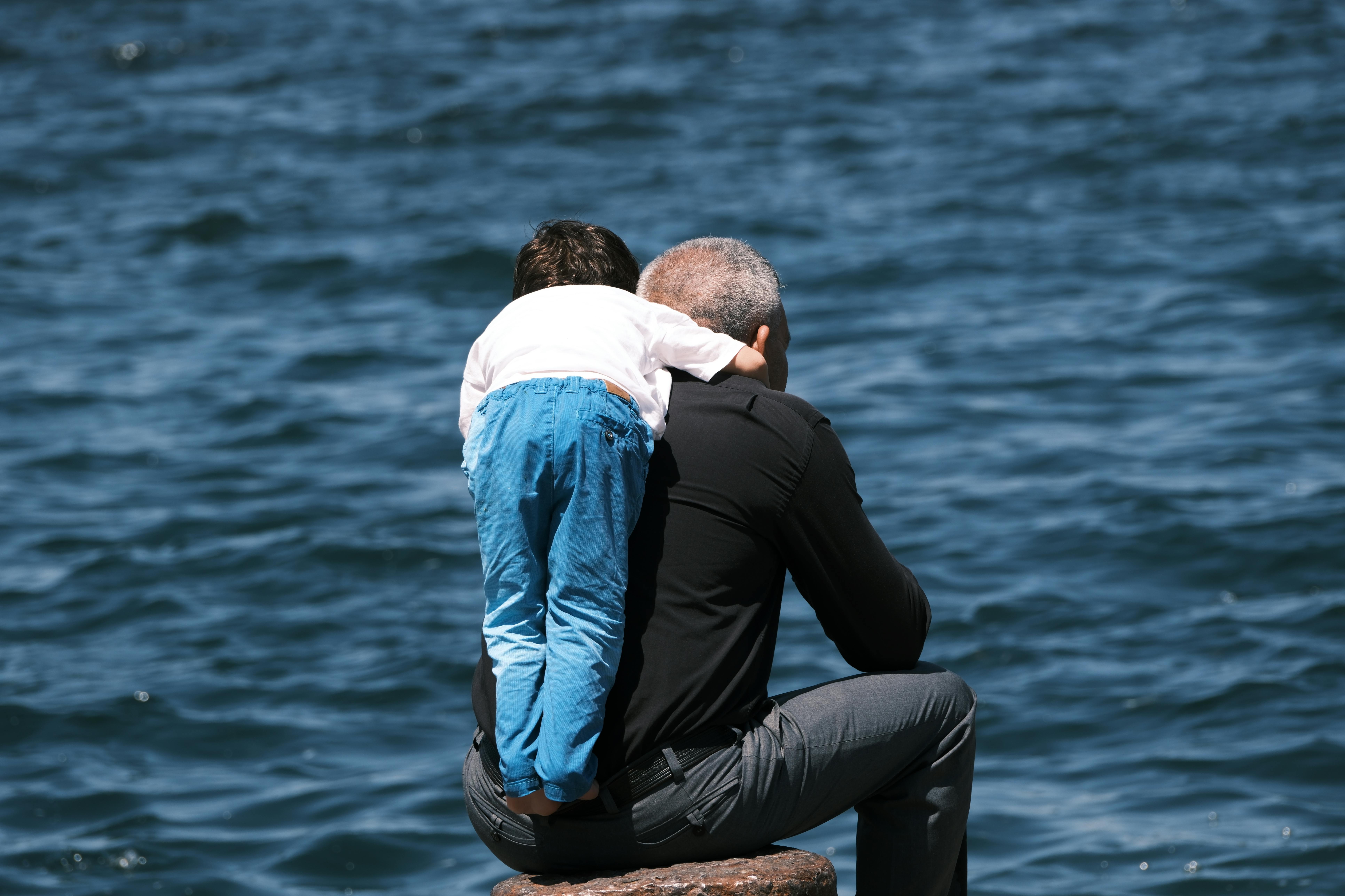 A little boy leaning on Grandfather shoulder sitting on the river bank