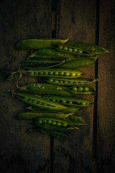 A dark, moody image of fresh green peas in pods arranged on a rustic wooden background.