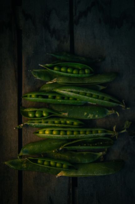 A pile of shelled green peas next to a few open pods on a wooden cutting board.