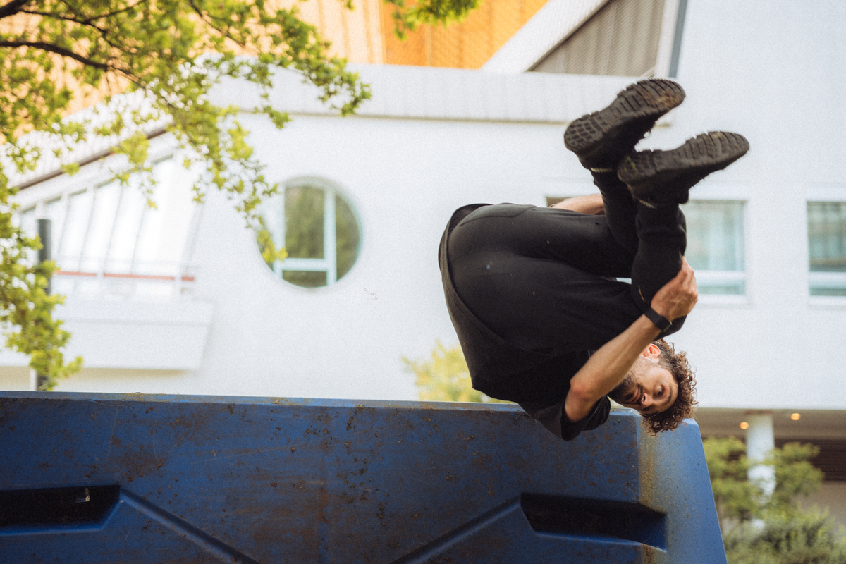 A freerunner fully inverted mid-flip above a blue parkour obstacle in a park.