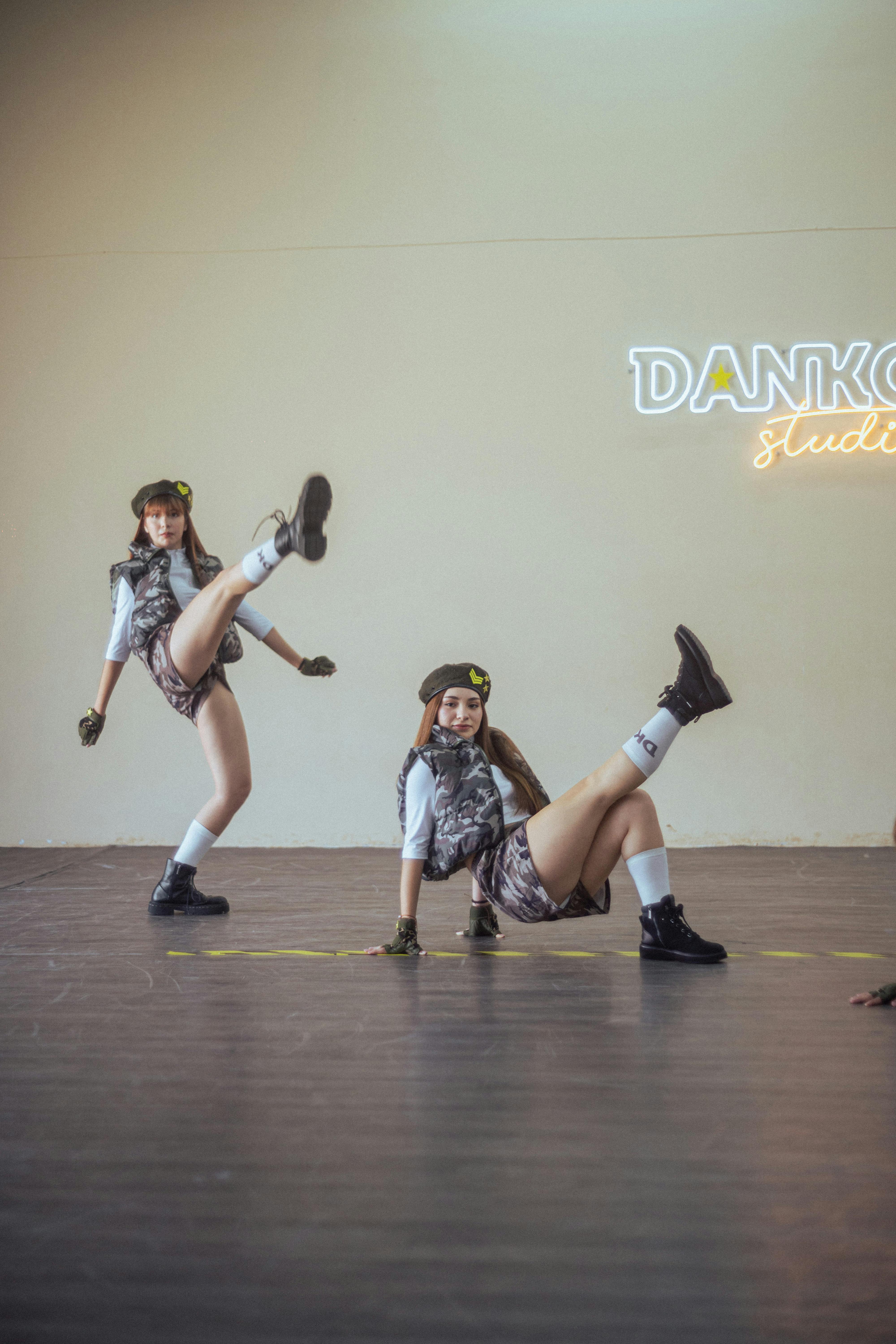 Two female dancers in military-inspired outfits perform dynamic moves in a studio setting.