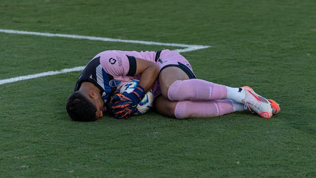A soccer player in pink gear resting on the field, hugging a ball, possibly taking a break after a match.