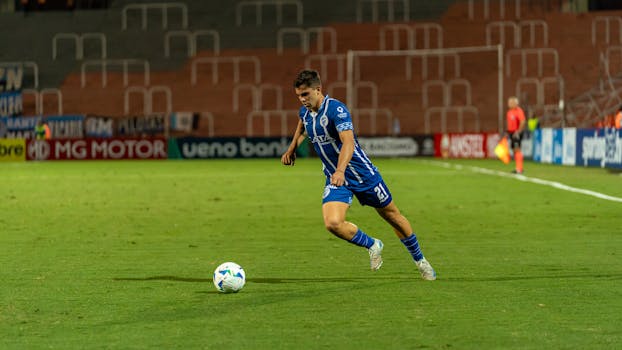 A soccer player in blue jersey dribbling the ball on a well-lit field during a night match.