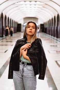 Woman in leather jacket poses confidently in modern subway corridor.