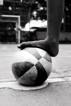 Black and white photo of a foot balancing on a soccer ball on an outdoor court.