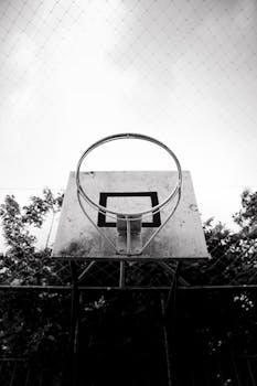 Black and white photo of a basketball hoop against a cloudy sky, creating a dramatic and nostalgic atmosphere.