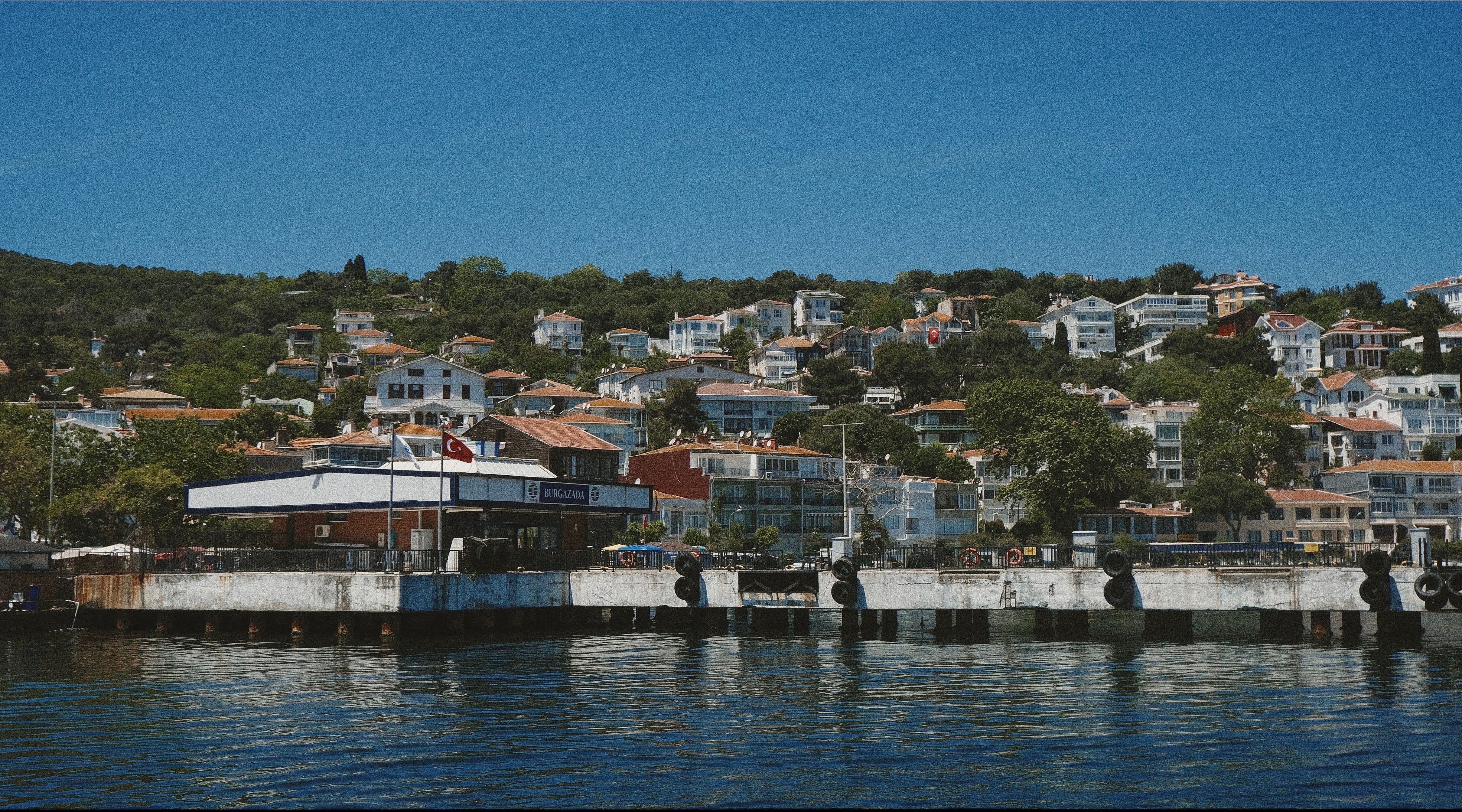 Ferry station with hillside homes in Burgazada, Istanbul, under a clear blue sky.