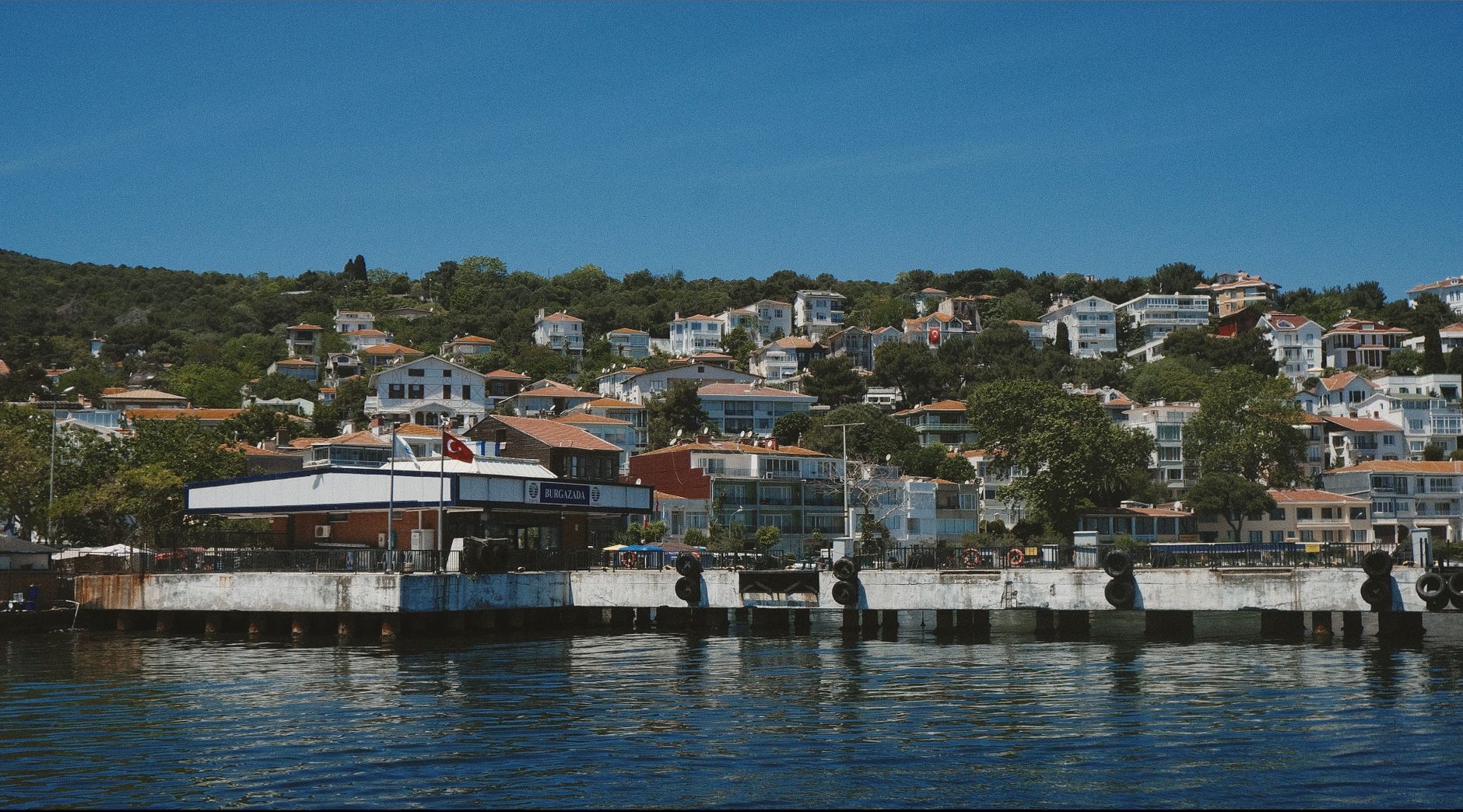 Ferry station with hillside homes in Burgazada, Istanbul, under a clear blue sky.