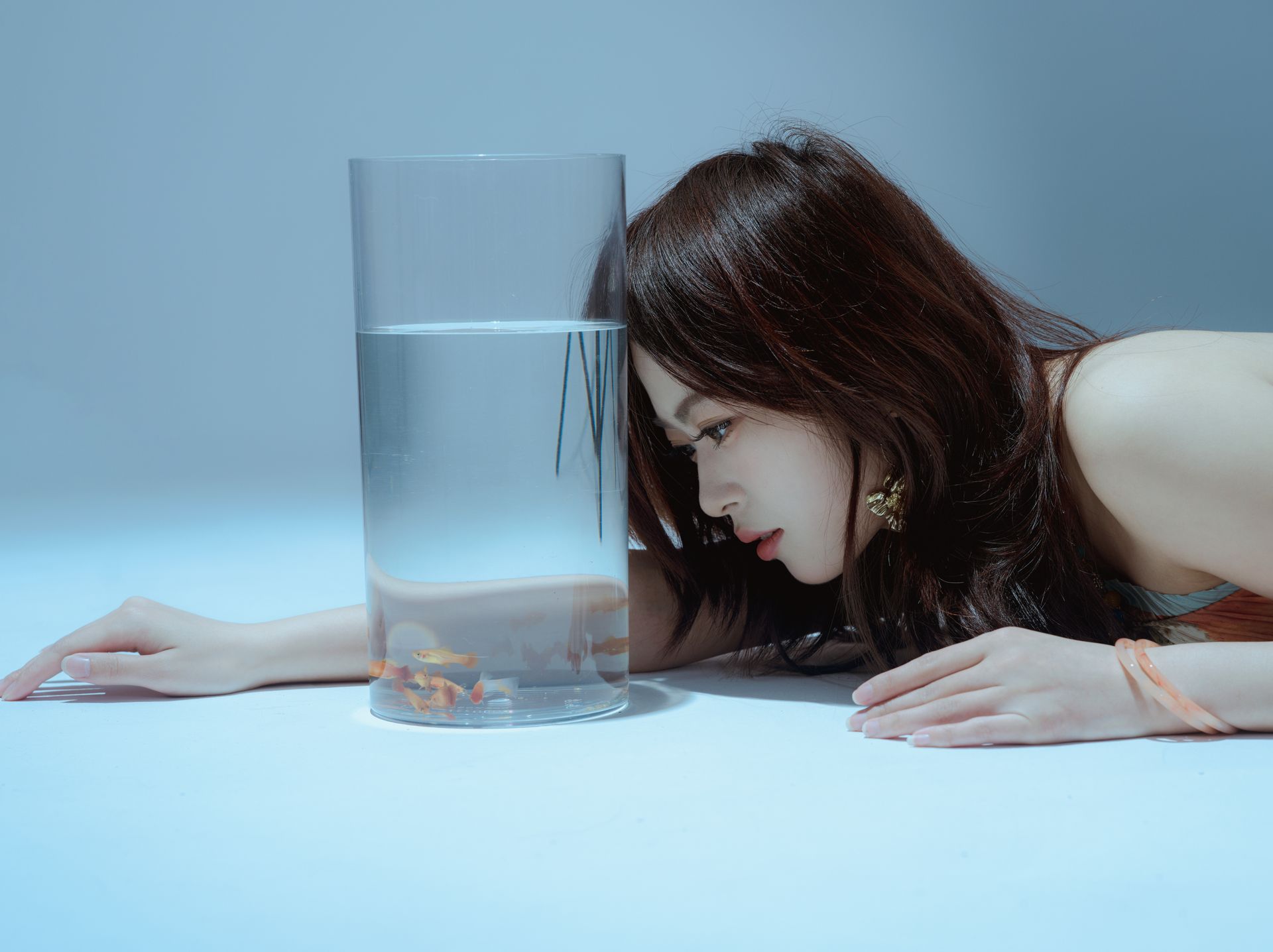 A woman intently observes goldfish swimming in a clear vase on a minimalist white surface.
