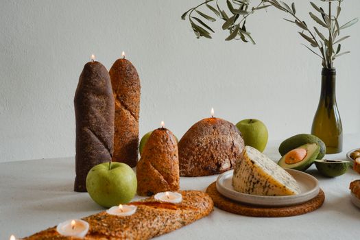 A rustic tabletop with bread-shaped candles, cheese, apples, and avocados, styled with leafy branches.
