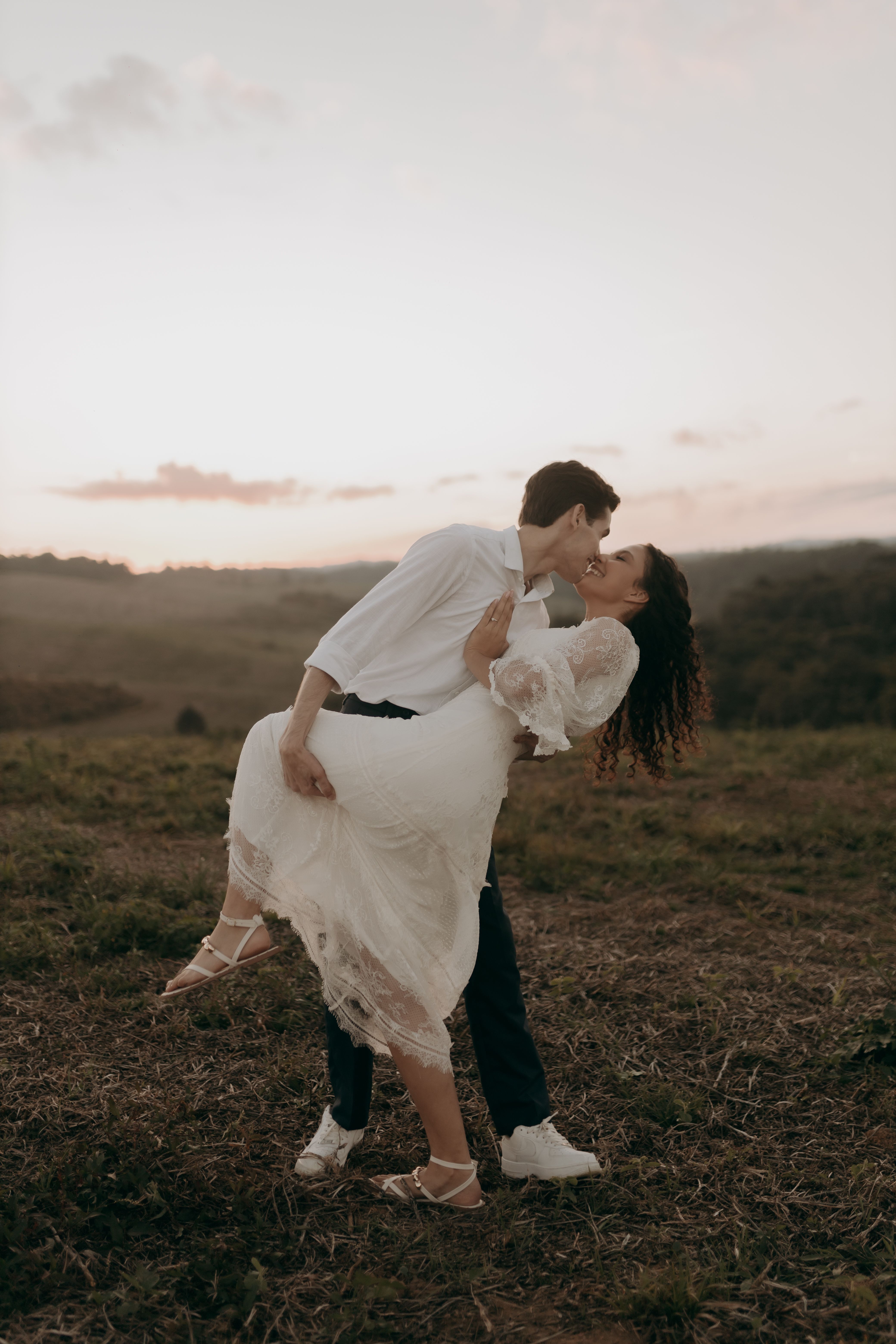 A couple in formal attire shares a romantic kiss during a sunset in a beautiful natural setting.