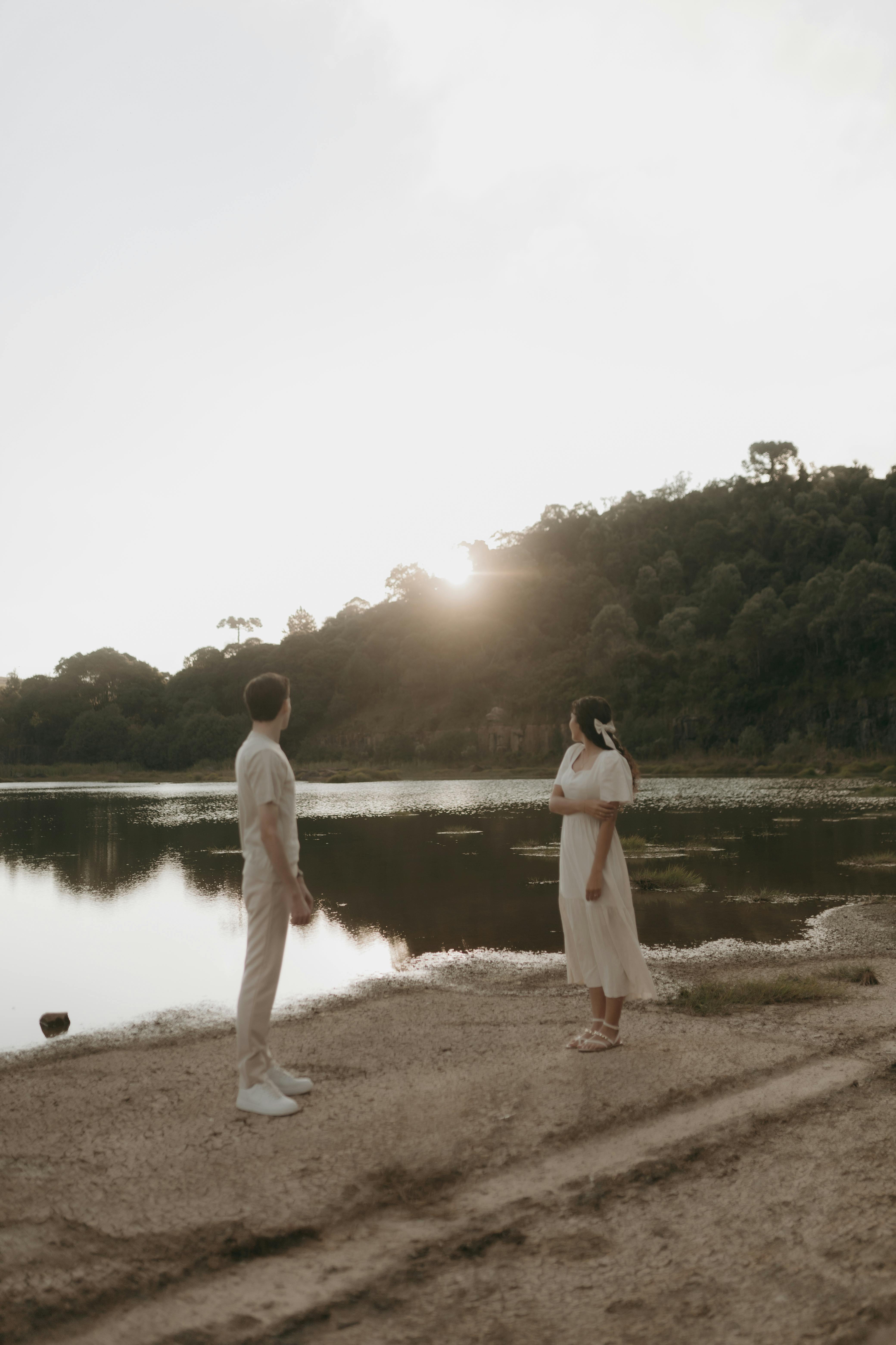 A couple standing by a serene lake during sunset, surrounded by nature's tranquility.