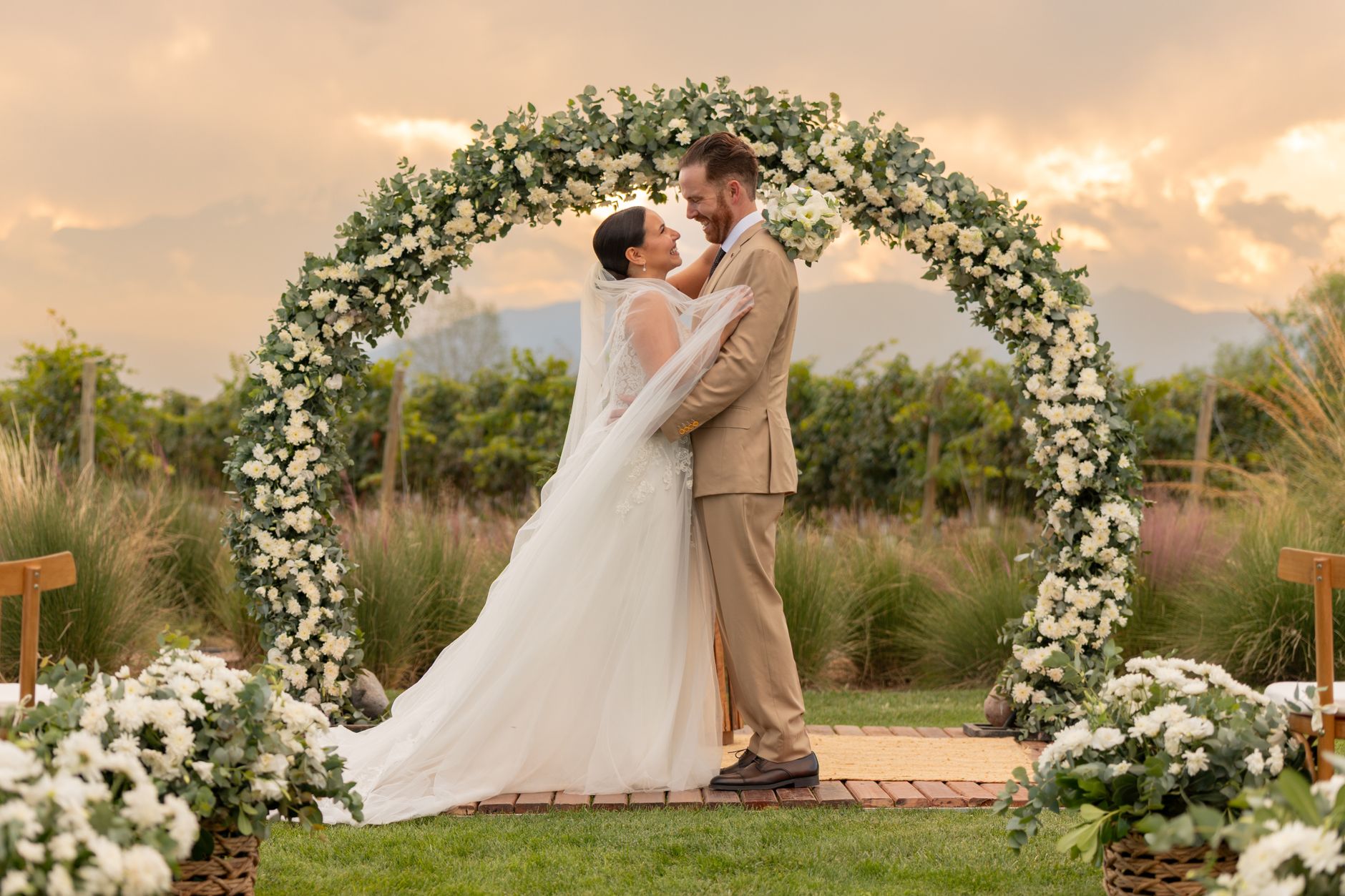 Bride and groom embrace under floral arch in Mendoza vineyard at sunset.