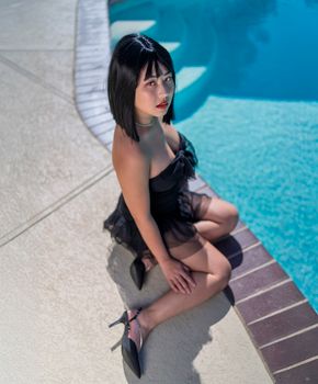 Elegant woman in a black dress sitting by a pool, showcasing modern summer fashion.