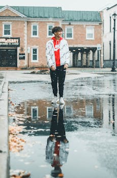 一名男子穿着时尚服装，走在潮湿的城市街道上，倒影映在水坑中