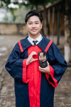 A graduate in gown holds a smartphone displaying a video call outdoors.