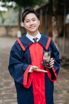 Smiling graduate in red and blue gown showing phone outdoors.
