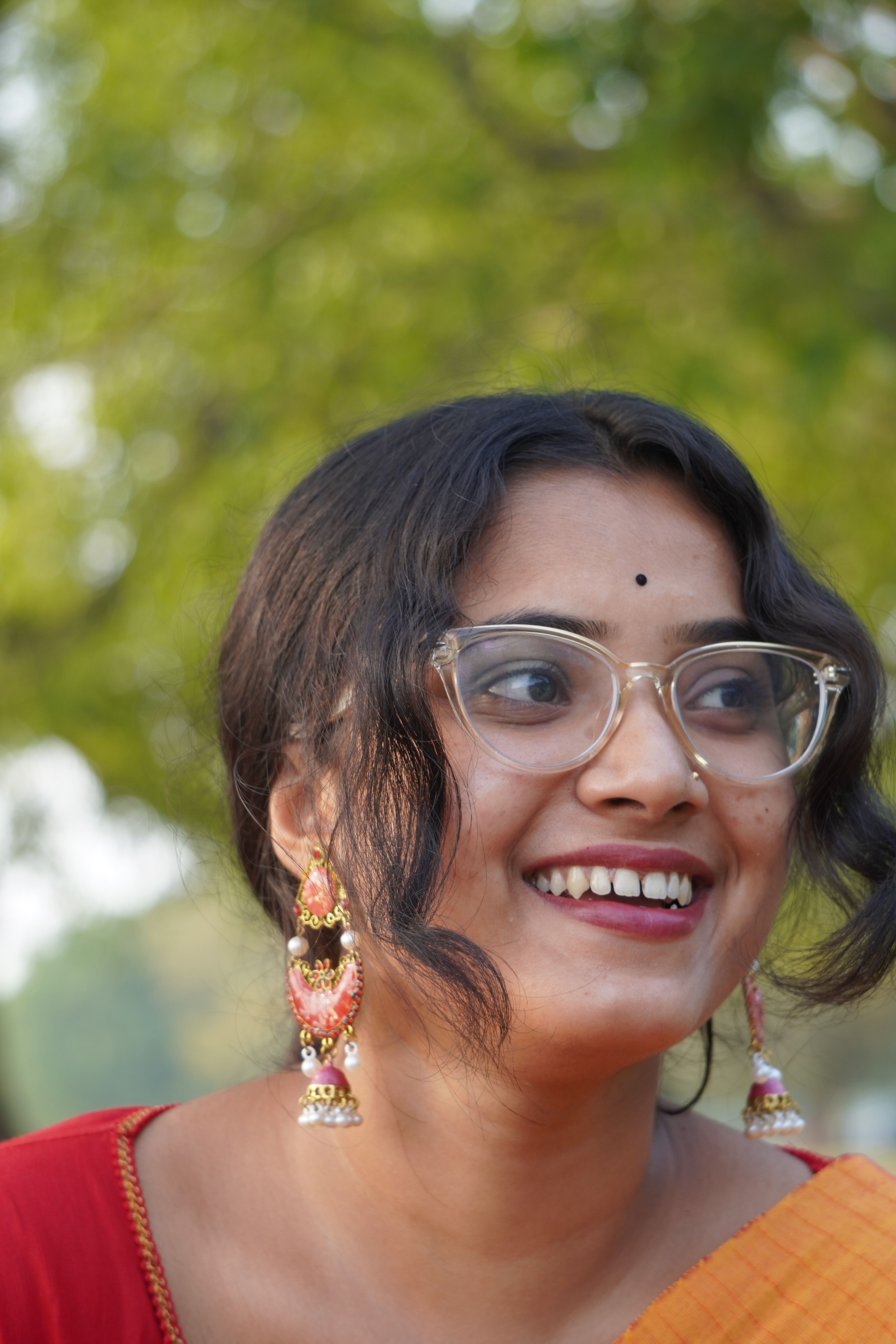 A young woman in stylish glasses smiles while wearing traditional attire, set against a natural outdoor backdrop.