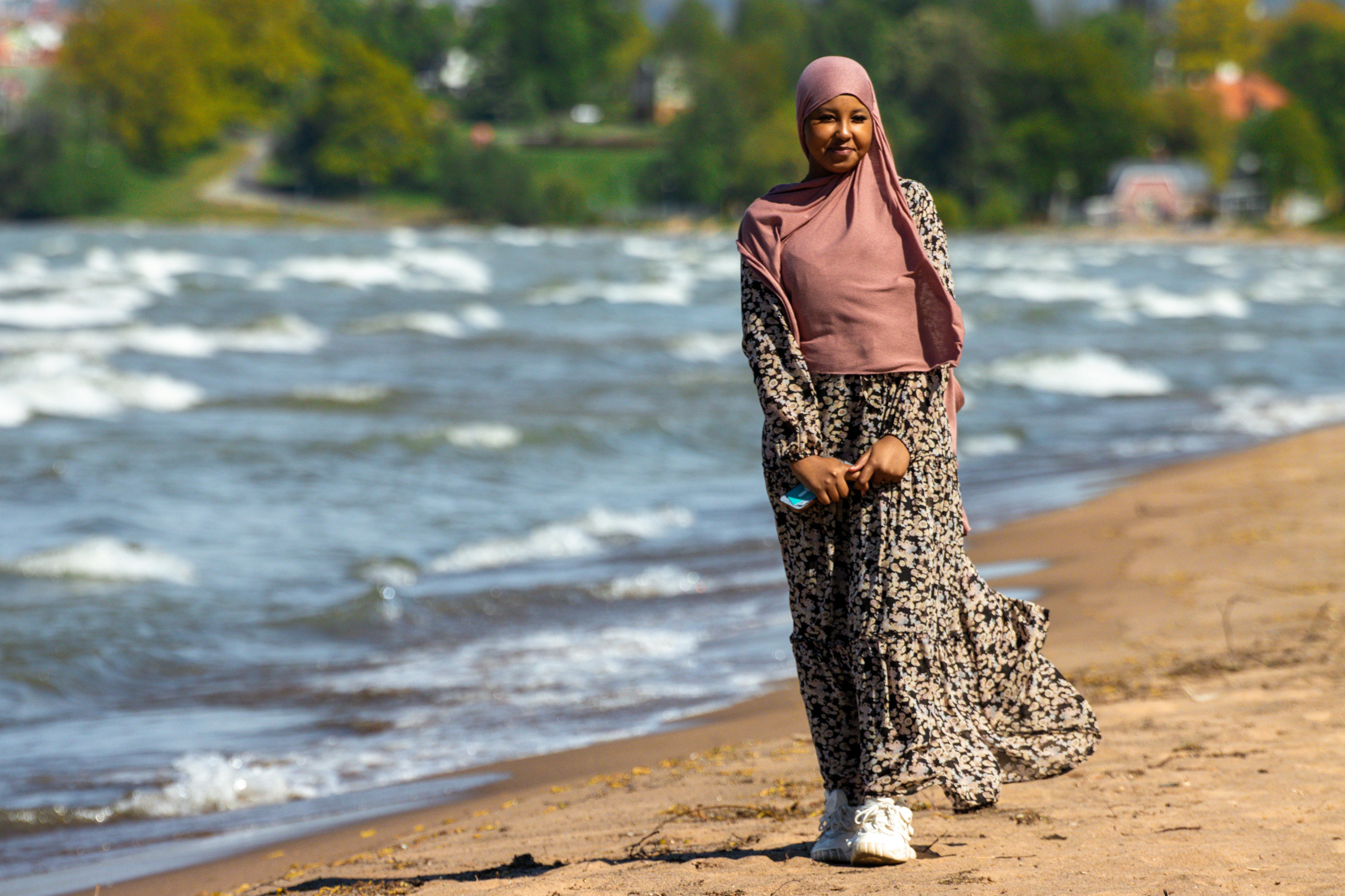 A woman in a hijab strolls along the shore of a beach in Jönköping, Sweden, on a sunny day.