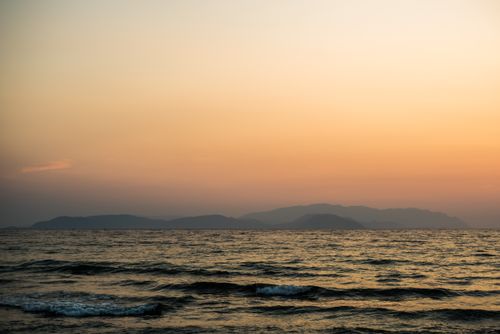 Free A tranquil ocean view at sunset with gentle waves and distant mountains under an orange sky. Stock Photo