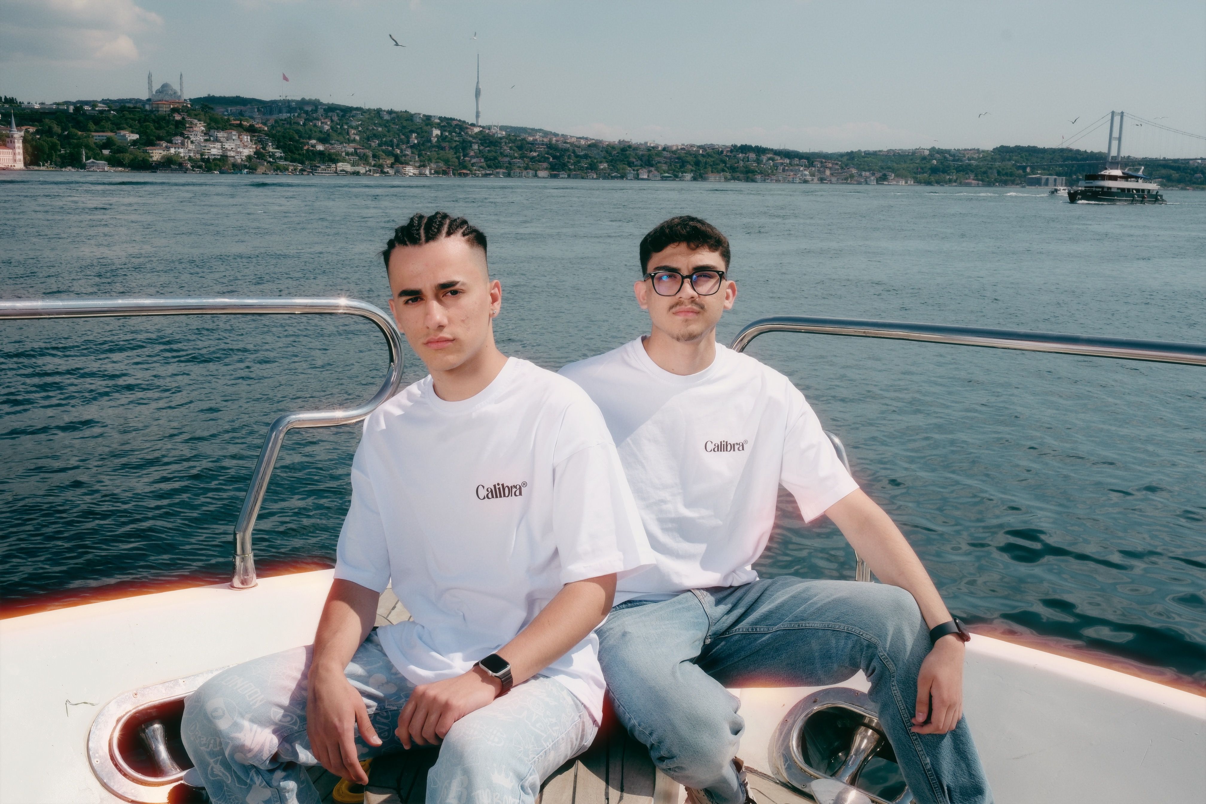 Young men in trendy attire pose on a boat with the Bosphorus Bridge in the background.