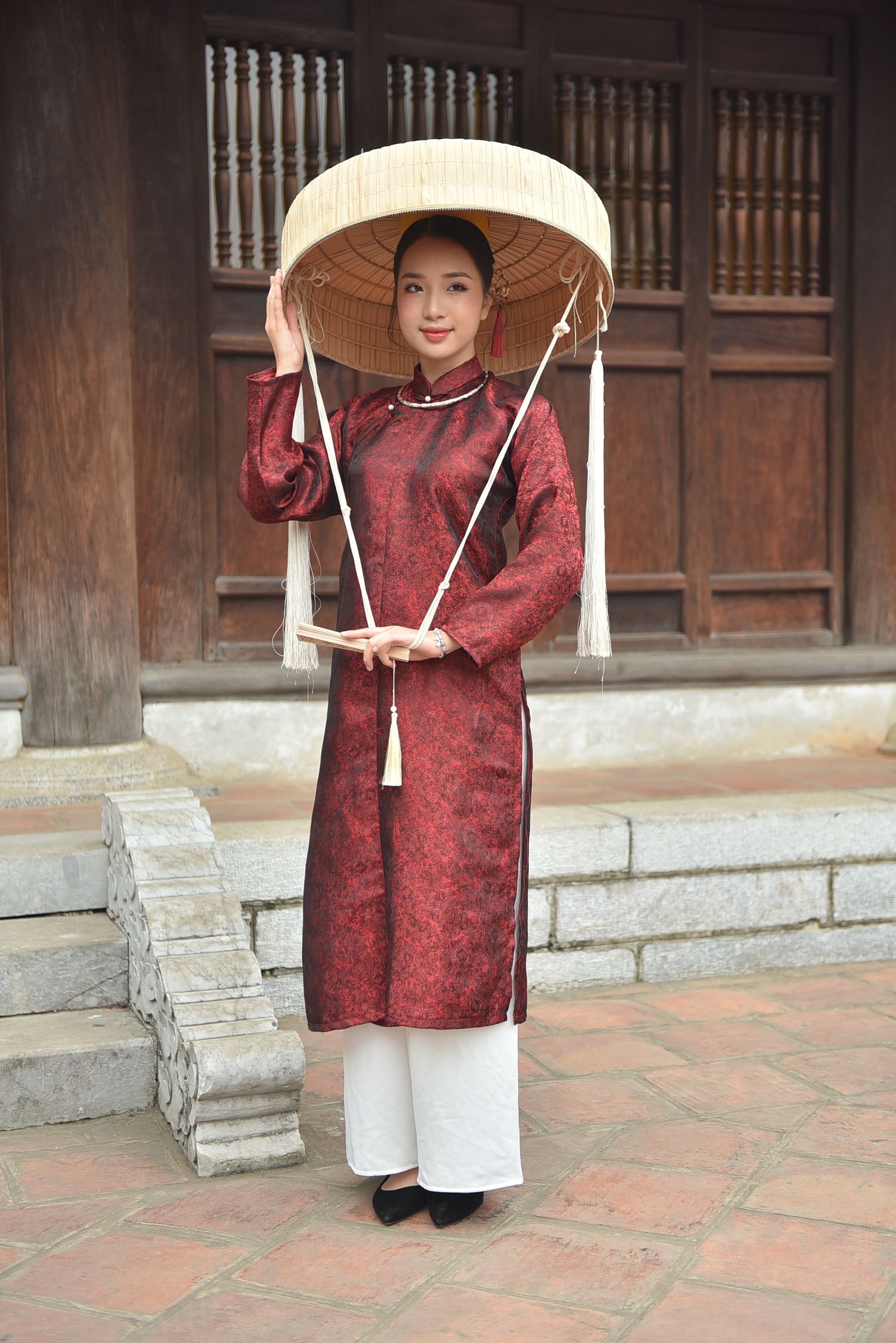 Người phụ nữ Việt Nam diện áo dài truyền thống kèm nón lá đứng trước căn nhà gỗ cổ truyền
