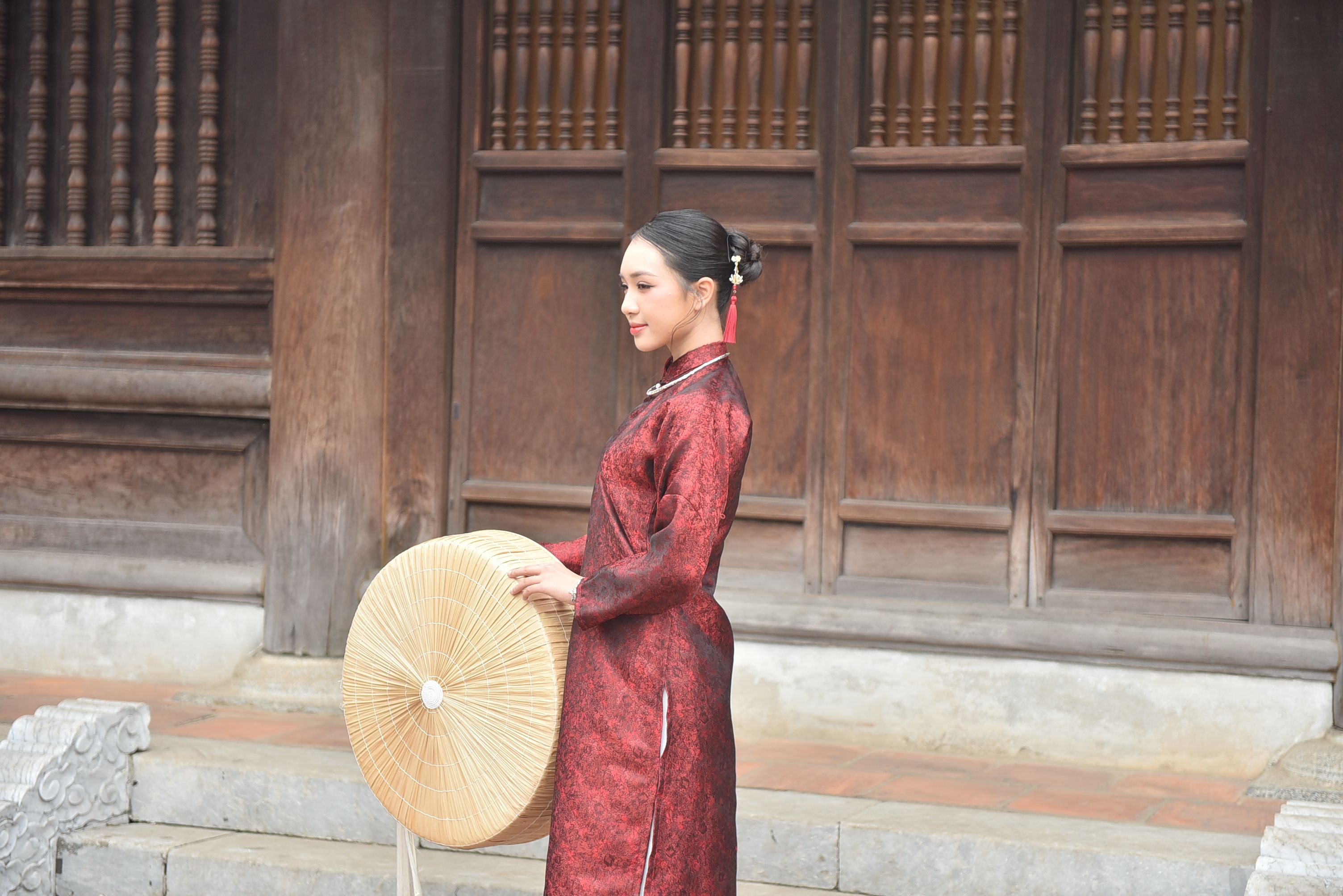 Woman in a red traditional dress holding a straw basket standing outside a wooden building.