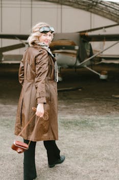Stylish woman in aviator goggles stands by a vintage aircraft, embracing retro fashion.