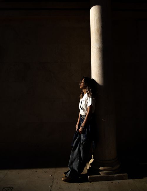 Free A woman stands against a column in a play of light and shadow outdoors. Stock Photo