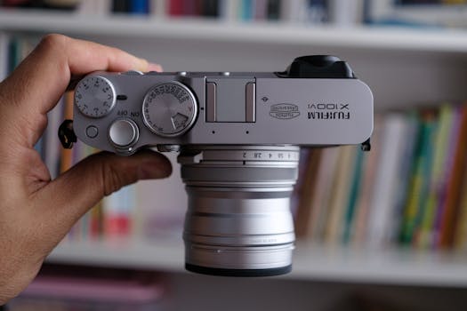 Hand holding a silver Fujifilm X100V camera with control dials displayed clearly.