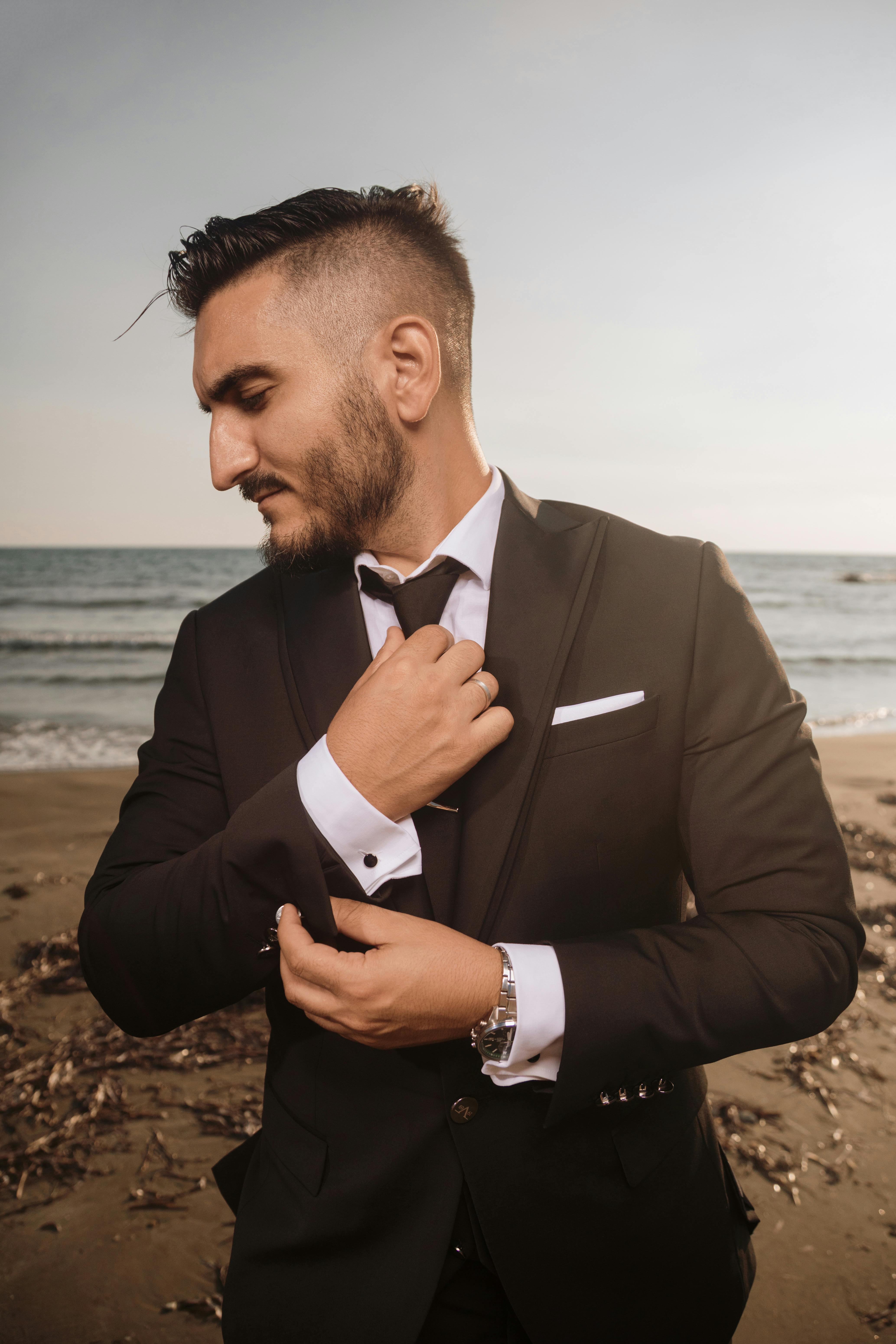 Confident man in a stylish suit adjusting his cufflinks on a beach during sunset, exuding elegance.