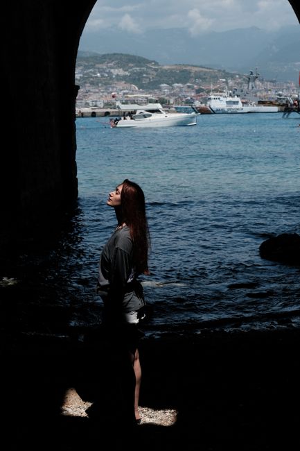 Woman silhouetted under archway by sea with yachts and cityscape.