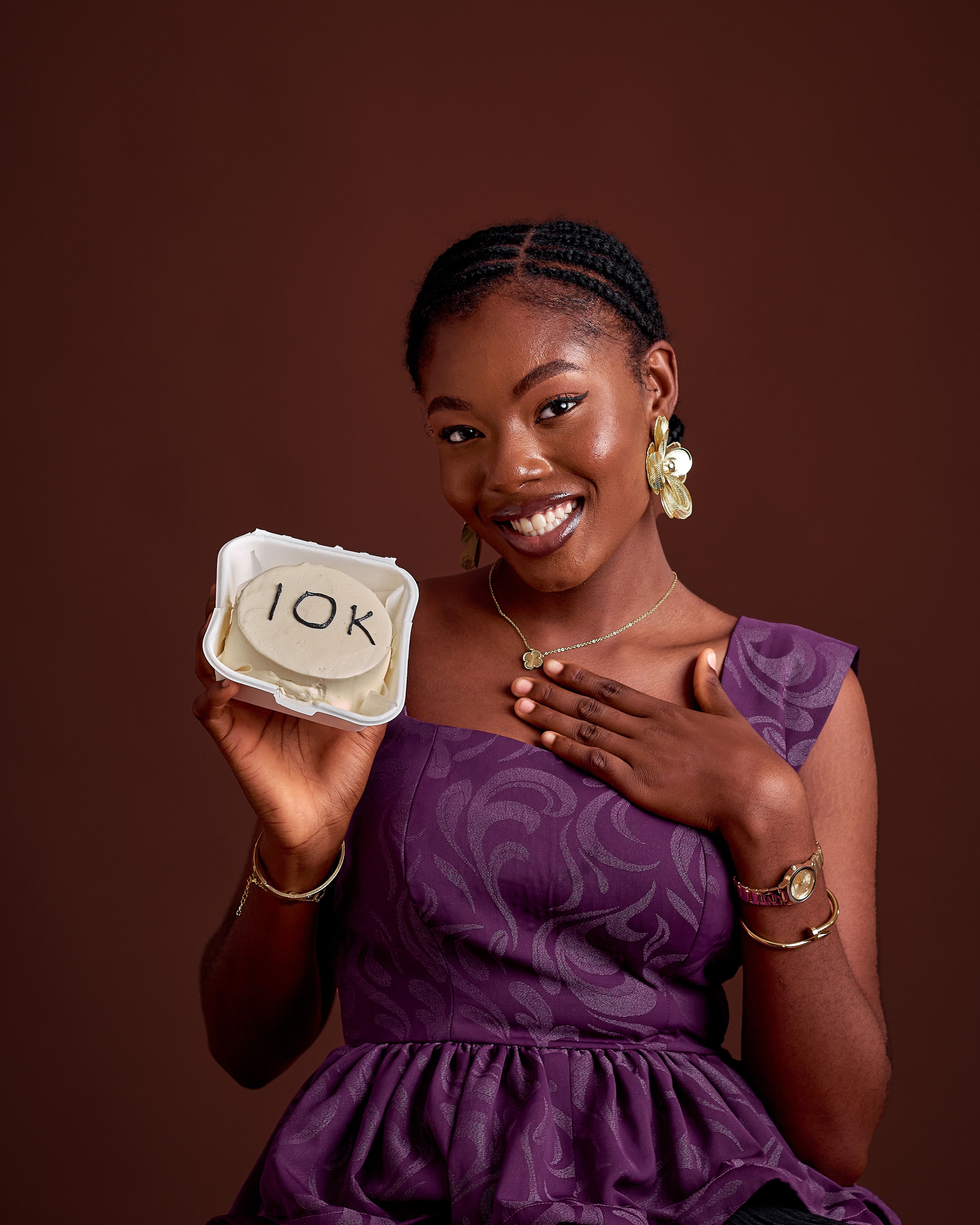 African woman in elegant dress smiling, holding a 10K inscribed cake.