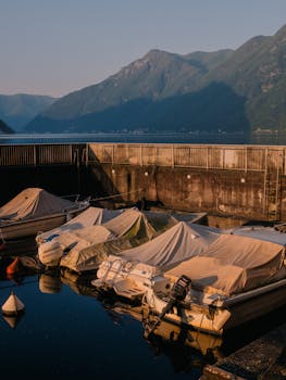Covered boats docked by a tranquil lake surrounded by mountains at dawn.