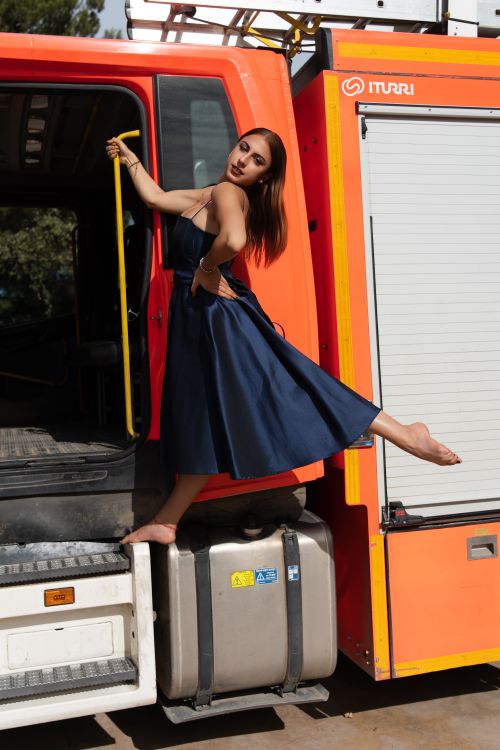 Free Confident woman in blue dress posing playfully on a fire truck step outdoors. Stock Photo