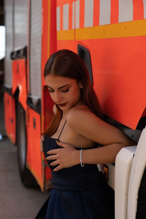 Free A woman in a dark dress poses by a vibrant orange fire truck, capturing a mix of elegance and service. Stock Photo