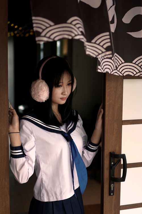 Free Young woman wearing a sailor uniform and earmuffs, standing at a doorway with cultural decor. Stock Photo