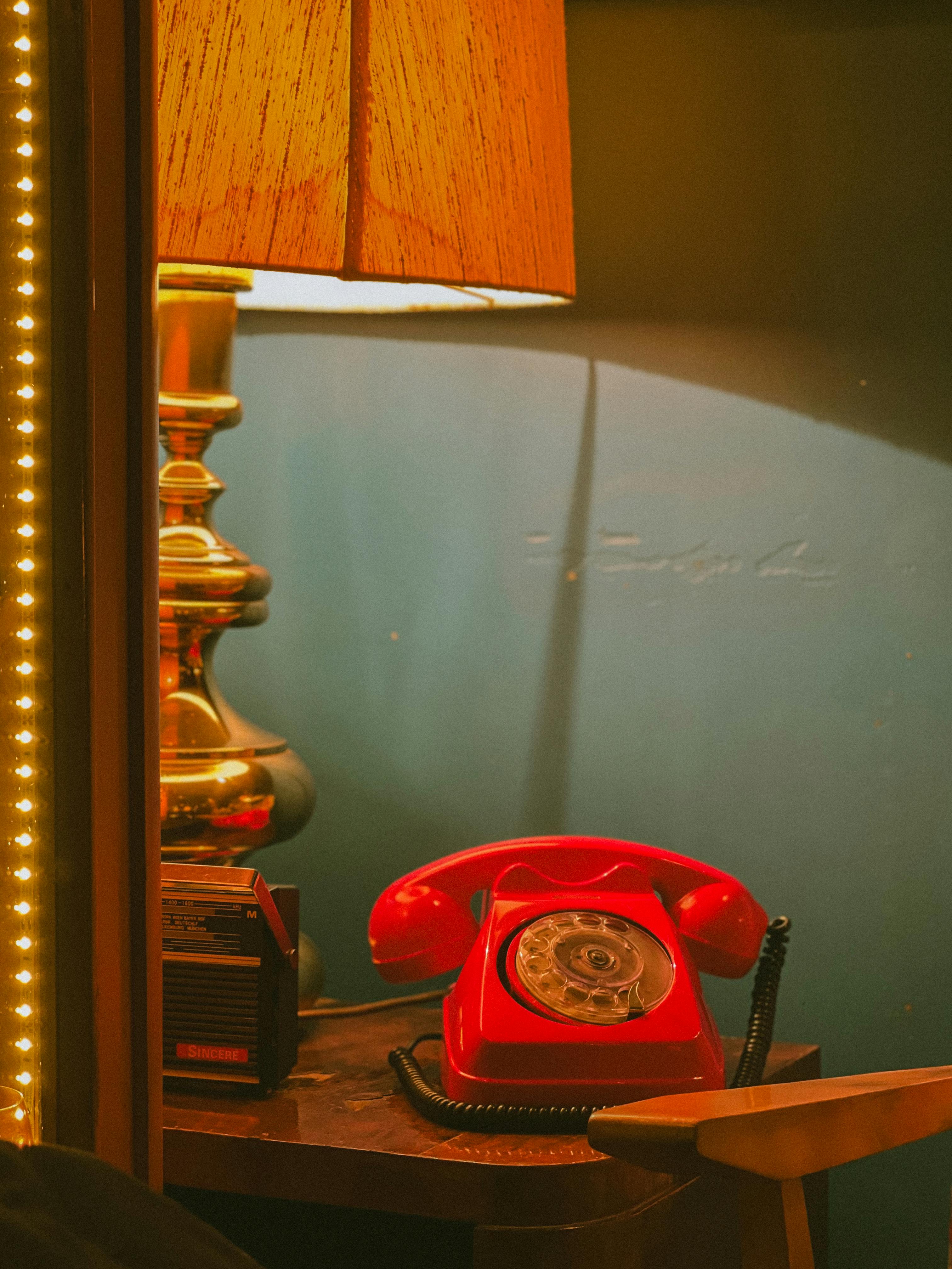 Kostenlos Eine nostalgische Umgebung mit einem roten Wählscheibentelefon, einer Tischlampe und einem klassischen Radio auf einem Holztisch. Stock-Foto